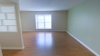 This single-floor space presents a clean, open layout designed to maximize usability and comfort. The main living area is defined by smooth, polished hardwood flooring that reflects natural light, lending the space a warm and inviting ambiance. The floors are well-maintained and provide a durable, attractive foundation throughout.

The walls are painted in a neutral tan tone along the main wall, transitioning to a soft sage green accent wall, creating a subtle yet refreshing contrast. White baseboards run along the perimeter, adding a crisp finishing detail that complements the overall color palette.

A centrally placed window along the back wall allows natural light to filter into the room, brightening the interior and contributing to an airy feel. The smooth, white ceiling enhances the sense of openness within the space.

The room's generous proportions allow for flexible furniture arrangements, making it well-suited for use as a living room, lounge, or multi-purpose area. Overall, the space combines practical design elements with a neutral aesthetic, providing a versatile and move-in ready environment adaptable to a variety of uses.