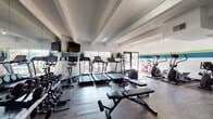 Located at 11100 E Dartmouth Ave in Denver, Colorado, this well-equipped fitness center is designed to support a comprehensive workout experience on a single level. The space is defined by its clean, modern aesthetic, featuring white walls accented by bold horizontal stripes in teal and green that run throughout, giving the room a vibrant, energetic atmosphere. Dark wood-look flooring grounds the space and contrasts effectively with the bright interior.

The cardio zone is well-appointed with multiple treadmills, elliptical trainers, and stationary spin bikes, all positioned to take advantage of large sliding glass windows that offer views of the outdoor grounds and pool area, allowing natural light to fill the space. Wall-mounted flat-screen televisions provide entertainment during workouts.

A dedicated strength training area houses a cable cross machine, a multi-function cable station with digital instructional kiosks, and additional weight machines. Adjustable benches and a full rack of dumbbells round out the free weights section, catering to a variety of training needs. Full-length mirrors are mounted along one wall, enhancing the sense of openness and allowing users to monitor their form. Track lighting illuminates the entire space efficiently, and a textured ceiling with exposed beams adds a subtle architectural character to the room.