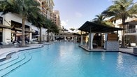 This expansive outdoor pool area exudes a luxurious resort atmosphere, centered around a large, crystal-clear lagoon-style pool with vivid turquoise water. The pool's generous size and smooth surface create a striking visual focal point, reflecting the surrounding architecture and open sky. Flanking the water's edge are elegantly designed private cabanas featuring sleek, modern canopies draped with flowing white curtains, offering shaded retreats with seating for relaxation and entertaining. The cabanas extend directly over or alongside the pool, enhancing the sense of exclusivity and seamless indoor-outdoor living. Lush, mature palm trees are thoughtfully distributed throughout the space, adding a tropical ambiance and natural greenery that complements the resort aesthetic. Sun loungers and poolside seating are arranged along the deck areas, providing ample space for leisure. The surrounding multi-story building features warm-toned facades and private balconies overlooking the pool, creating an intimate yet grand setting. The overall ambiance is polished and sophisticated, blending tropical landscaping with upscale architectural details to deliver a premier outdoor living and leisure experience.