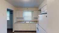 This single-floor residence offers a practical and cohesive layout across its living spaces. The kitchen serves as a functional centerpiece, equipped with white shaker-style cabinetry both above and below granite-style countertops. A stainless steel sink sits beneath a window fitted with horizontal blinds, allowing natural light to brighten the workspace. White appliances, including an electric range with overhead ventilation, a refrigerator, and a microwave, complete the kitchen's setup. Light wood-style flooring runs throughout the kitchen, contributing to a clean, bright aesthetic.

The living area features wall-to-wall carpeting in a neutral tone, with a window unit providing climate control and a baseboard heating system along the walls. A doorway connects the living space directly to the kitchen, supporting an open flow between areas.

The bedrooms are carpeted and finished with neutral walls. Each room includes windows with blinds that invite natural light, and one bedroom benefits from two windows on adjacent walls. Baseboard heaters are present for supplemental warmth.

The bathroom is straightforward and cleanly finished, featuring a full-size soaking tub with a showerhead and white square tile surround, complemented by light flooring and neutral walls throughout.