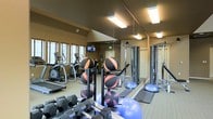 This fitness center offers a comprehensive workout environment designed to accommodate a wide range of exercise needs. The space features vaulted ceilings that enhance the sense of openness, while large windows along one wall allow natural light to filter in, creating an inviting atmosphere. Overhead fluorescent lighting ensures the room remains well-lit throughout the day.

The cardio section includes multiple treadmills and elliptical machines arranged along the windowed wall, complemented by a wall-mounted television for entertainment during workouts. A full-length mirror runs along one side of the room, visually expanding the space while allowing users to monitor their form.

The free weights area is well-stocked with a range of hex dumbbells neatly arranged on a rack, alongside a medicine ball tower holding multiple weighted balls. Balance and flexibility training is supported by the presence of BOSU balls and exercise mats. A multi-functional cable and strength training machine anchors the far end of the room, offering versatile resistance training options. The neutral-toned walls and carpeted flooring contribute to a clean, functional aesthetic throughout the space.