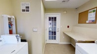 This single-floor utility space is designed with functionality in mind. The room features light beige walls and neutral carpeted flooring, creating a clean and practical environment. A washer unit is positioned along one wall, complemented by a built-in countertop that extends along the adjacent wall, offering ample workspace for folding laundry or other tasks.

A white-framed exterior door with a multi-pane glass window serves as a prominent feature, allowing natural light to filter into the space while providing direct outdoor access. The glazed door offers views to the outside, adding a sense of openness to the otherwise utilitarian room.

Overhead, a flush ceiling light ensures consistent illumination throughout the space. HVAC vents are present on both the wall and ceiling, ensuring proper climate control and ventilation. A security panel and fire alarm are mounted on the walls, highlighting the safety features integrated into the space. The overall layout is efficient and straightforward, making it a highly practical utility room suited for everyday household tasks.