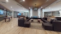Located at 201 E Mississippi Ave in Denver, CO, this single-floor amenity space offers a sophisticated, open-concept layout designed for both relaxation and social gathering. Wide-plank hardwood flooring flows throughout the main area, anchored by a striking double-sided linear fireplace set within a tall textured column with integrated open shelving. Multiple seating arrangements with plush modular sofas and mid-century accent chairs create distinct lounging zones beneath a warm wood-paneled ceiling and statement chandelier lighting. A dedicated media lounge features a large wall-mounted flat-screen TV set against rich wood paneling, complemented by leather armchairs, upholstered ottomans, and patterned carpet tiles for an intimate viewing experience. A wet bar with marble countertops and under-counter refrigeration connects seamlessly to the media area. The expansive chef's kitchen showcases a dramatic oversized island with a marble surface and leather bar stools, paired with two-tone cabinetry in warm wood and matte black finishes, a tile backsplash, stainless steel appliances, and globe pendant lighting. A billiards table and additional lounge seating round out the game and social zone, making this a comprehensive, resort-style amenity space.