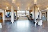 Located at 1390 Fox Valley Center in Aurora, Illinois, this striking lobby space offers an impressive first impression through its grand revolving entrance door, framed by floor-to-ceiling glass that floods the interior with abundant natural light and provides views of the surrounding exterior. The expansive single-floor layout is finished throughout with large-format grey marble-effect tiles that lend a polished, sophisticated character to the entire space.

Towering ceilings define the architectural identity of the lobby, supported by substantial cylindrical columns with a smooth, neutral finish. Cascading pendant light fixtures with gold accents punctuate the ceiling, complementing a dramatic chandelier installation near the reception area. A bold teal accent wall adorned with a sculptural bronze butterfly art piece serves as a striking focal point, further elevated by a cascading globe light installation overhead.

The space is thoughtfully divided into distinct zones: a welcoming reception desk with a curved, stone-clad design; comfortable lounge seating areas featuring modern upholstered chairs and sofas in teal, plum, and neutral tones; and a wood-slat partition wall that adds warmth and visual texture. A wide central staircase with gold railings leads to an upper mezzanine level, enhancing the sense of vertical grandeur. Large windows throughout ensure the space remains bright and connected to the outdoors.