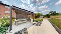 Located at 1803 Cobourg Court in Parkville, Maryland, this community's outdoor grounds offer a well-appointed collection of amenities set against a backdrop of classic multi-story brick buildings. At the heart of the space is a modern covered pergola structure fitted with decorative string lighting, slatted privacy screens, and a built-in outdoor kitchen complete with a grill and prep station. Concrete bench seating beneath the pergola provides a comfortable gathering area for residents. Adjacent to the pergola, meticulously maintained garden beds feature vibrant red flowering plants, ornamental grasses, and decorative rock landscaping that add color and texture to the grounds. A dedicated children's playground, surfaced with protective wood chip mulch, offers climbing structures and slides, providing a safe recreational area for younger residents. Expansive stretches of open lawn extend across the property, bordered by mature trees that offer natural shade and greenery. The brick exterior buildings feature private balconies overlooking the landscaped courtyard. Concrete walkways and gravel paths connect the various outdoor areas, ensuring easy navigation throughout the community grounds.