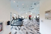 Located at 1700 Katy Fort Bend Road in Katy, TX, this well-appointed fitness center offers a generously sized, single-floor exercise space designed to accommodate a full range of workout needs. The room's most striking architectural feature is its soaring vaulted ceiling, which creates an open, airy atmosphere and is complemented by a ceiling fan for added comfort. Recessed lighting throughout ensures the space is well-lit at all times.

Large, black-framed windows line the walls, flooding the interior with natural light and providing views of the surrounding outdoor greenery. A full-wall mirror along one side of the room enhances the sense of space while providing functional feedback for exercise form.

The floor is covered in a distinctive chevron-patterned carpet in shades of gray and white, adding a modern, stylish aesthetic to the space. Equipment is thoughtfully arranged to maximize usability, and includes a row of TRUE brand treadmills, elliptical machines, stationary bikes, a cable and pulley functional training rig, TRX suspension trainers, a comprehensive dumbbell rack, and various strength machines. Additional accessories such as medicine balls, resistance tools, and storage shelving round out the facility. Warm wood-toned doors and built-in shelving add a refined touch to the overall design.