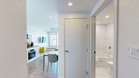 Located on the 54th floor in Calgary's vibrant urban core, this one-bedroom, one-bathroom unit delivers a modern, refined living experience with stunning panoramic views of the city skyline and surrounding landscape. The open-plan living and kitchen area is anchored by warm hardwood flooring and crisp white walls, creating a clean, contemporary aesthetic throughout. Floor-to-ceiling windows flood the living space with natural light while framing sweeping city views, complemented by a built-in window bench that invites residents to take in the scenery. The kitchen features white cabinetry, integrated appliances including a wall oven, and a compact dining area with a marble-top table. The bedroom continues the bright, minimalist theme with large windows showcasing iconic Calgary landmarks, a wall-mounted TV, and tasteful botanical artwork. The bathroom is finished in elegant marble-effect wall tiles, complete with a floating vanity, vessel sink, a large mirror, and a glass-enclosed shower. Recessed lighting throughout enhances the clean lines and polished atmosphere of this well-appointed high-rise unit.