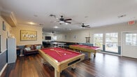 Located at 3200 S US Highway 1 in Fort Pierce, Florida, this single-floor recreational facility serves as a well-appointed community clubhouse designed for social gatherings and leisure activities. The space is finished with rich, dark hardwood-style flooring throughout, complemented by crown molding and a two-tone wall treatment featuring a coastal blue wainscoting panel paired with neutral upper walls — a nod to the surrounding Florida coastal environment.

The main game room is a standout feature, housing two full-size pool tables with red felt, a ping-pong table, and a shuffleboard table, all comfortably accommodated beneath multiple ceiling fans that provide excellent airflow. A lounge area with leather seating and a flat-screen TV creates a relaxed viewing and socializing zone. Natural light filters in through multiple windows with blinds, offering views of the surrounding greenery.

A secondary multipurpose room provides flexible space for tabletop games, crafts, or small group activities, equipped with folding tables, chairs, and a bookshelf stocked with books and board games. Beach-themed wall décor reinforces the cheerful, casual atmosphere throughout. The facility also includes two full bathrooms, a closet for storage, and additional unclassified utility spaces, making it a fully functional and welcoming recreational hub.