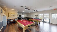 Located at 3200 S US Highway 1 in Fort Pierce, Florida, this single-floor recreational facility serves as a well-appointed community clubhouse designed for social gatherings and leisure activities. The space is finished with rich, dark hardwood-style flooring throughout, complemented by crown molding and a two-tone wall treatment featuring a coastal blue wainscoting panel paired with neutral upper walls — a nod to the surrounding Florida coastal environment.

The main game room is a standout feature, housing two full-size pool tables with red felt, a ping-pong table, and a shuffleboard table, all comfortably accommodated beneath multiple ceiling fans that provide excellent airflow. A lounge area with leather seating and a flat-screen TV creates a relaxed viewing and socializing zone. Natural light filters in through multiple windows with blinds, offering views of the surrounding greenery.

A secondary multipurpose room provides flexible space for tabletop games, crafts, or small group activities, equipped with folding tables, chairs, and a bookshelf stocked with books and board games. Beach-themed wall décor reinforces the cheerful, casual atmosphere throughout. The facility also includes two full bathrooms, a closet for storage, and additional unclassified utility spaces, making it a fully functional and welcoming recreational hub.