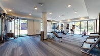 This modern fitness center is thoughtfully designed across a single floor, offering a comprehensive workout environment suited for a wide range of exercise routines. The space is divided into distinct functional zones, each finished with contemporary materials and quality fixtures throughout.

The main exercise area features striped, multi-tone carpeted flooring and is lined with large floor-to-ceiling windows that flood the space with abundant natural light and offer open views of the surrounding greenery. This zone is outfitted with a full array of cardio equipment, including treadmills, ellipticals, stationary bikes, and a rowing machine, all arranged to maximize both usability and sightlines to the outdoors. A multi-functional cable machine and adjustable weight bench anchor the strength training section, which is further enhanced by a full-length wall mirror that expands the sense of space.

A dedicated studio room provides a quieter setting for yoga, stretching, or group exercise. This area features smooth light wood flooring, a tray ceiling with sleek linear lighting fixtures, and modern wall art, contributing to a calm and focused atmosphere. A rack of stability balls and free weights lines one wall, keeping essentials within easy reach.

The facility also includes a clearly posted fitness rules display and a framed certification, reflecting a commitment to safety and professionalism. The overall design balances functionality with a clean, modern aesthetic.