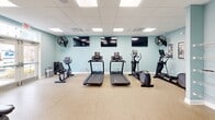 Located in Rehoboth Beach, Delaware, this fitness center offers a comprehensive workout environment designed to support a wide range of exercise routines. The space is finished with soft aqua-toned walls and durable speckled flooring, creating a clean and energizing atmosphere throughout.

The cardio zone features multiple treadmills, an elliptical trainer, and a stationary bike, all positioned beneath a row of wall-mounted flat-screen TVs and flanked by wall-mounted fans to maintain airflow during workouts. A full-wall mirror in the cardio area further enhances the sense of openness.

The strength training section is well-stocked with a tiered dumbbell rack, kettlebells, medicine balls, stability balls, and a multi-function cable machine with an adjustable bench — providing options for both free weight and machine-based training. A dedicated wall rack offers additional storage for accessories.

Practical amenities include built-in water fountains mounted to the wall for convenient hydration, as well as informational signage. Natural light filters in through windows on one side of the room, complemented by recessed panel lighting in the drop ceiling above. The layout is thoughtfully organized to allow ample space for movement between equipment stations, making this fitness center a fully functional and comfortable facility for daily exercise.