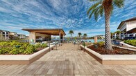 Located at 5400 Coraci Blvd in Port Orange, Florida, this expansive resort-style amenity complex delivers an impressive outdoor living experience spanning a generous footprint. The centerpiece is a large, sparkling swimming pool framed by natural stone decking and an abundance of blue lounge chairs, all set against panoramic waterfront views with a decorative fountain visible in the distance. Towering palm trees punctuate the landscape, reinforcing the tropical atmosphere throughout.

A prominent covered pavilion serves as a social hub, featuring a warm wood-paneled ceiling, ceiling fan, mounted flat-screen television, and open-air seating that overlooks both the pool and the waterway beyond. Beneath the pavilion, a fully equipped outdoor kitchen island with granite countertops and a stainless steel sink provides an ideal space for entertaining. Adjacent to this is a professional-grade stainless steel grilling station complete with a commercial hood vent and ample counter space.

The main building presents Mediterranean-inspired architecture, with arched windows, stone accents, and a pitched roof. Broad paver staircases with wrought iron railings lead to the building entrance, where floor-to-ceiling glass walls flood the interior with natural light. Waterfront lounge areas along the stone-paved perimeter offer serene spots to relax with unobstructed lake views.