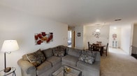 This single-floor space features an open-concept layout that seamlessly connects the living and dining areas, creating a functional and cohesive environment. The living area is anchored by a generously sized grey sectional sofa accented with decorative patterned pillows, complemented by a glass-top coffee table. A vibrant floral canvas artwork in red and orange tones adds a pop of color to the otherwise neutral white walls, contributing warmth to the space. The flooring transitions between carpet in the living zone and what appears to be a harder surface near the dining area. The dining space is defined by a dark wood table paired with matching chairs, illuminated by an elegant branching chandelier that serves as a decorative focal point. A doorway visible from the living area leads to a bedroom, suggesting a straightforward and accessible floor plan. The overall ambiance is casual and practical, with neutral tones throughout the walls and ceiling providing a clean, understated backdrop. Recessed and pendant lighting ensure the space is well-lit, making it feel inviting and comfortable for everyday living.