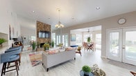 This single-floor clubhouse offers an open-concept layout designed for gathering and socializing. The main living area serves as the centerpiece of the space, anchored by a dramatic floor-to-ceiling natural stone fireplace with a wall-mounted television above. Soaring ceilings enhance the sense of openness, while a chandelier adds a refined touch to the seating area, which features comfortable furnishings arranged over vibrant, colorful area rugs atop light wood-style flooring that runs throughout the space.

A built-in bar counter with stylish blue barstools provides a convenient service area, adding functionality to the clubhouse setting. Adjacent to the main lounge, a dedicated dining area is bathed in natural light from large windows with views of the surrounding greenery. A ceiling fan overhead ensures year-round comfort in this bright, windowed nook furnished with a round dining table and patterned upholstered chairs.

French doors at the rear of the space open to the outdoors, further connecting the interior to the surrounding environment. The overall ambiance is warm and inviting, blending natural textures — such as the stone fireplace surround — with light, neutral walls and eclectic décor accents that create a welcoming community atmosphere.