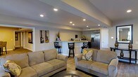 This single-floor space offers a thoughtfully designed open-concept layout that seamlessly blends the living area with a functional kitchen bar. The living room is anchored by two plush, neutral-toned sofas arranged to foster comfortable conversation, complemented by a glass-top coffee table that adds a touch of sophistication. Recessed lighting is installed throughout the sloped ceiling, providing bright, even illumination across the entire space. The kitchen area features a raised breakfast bar with three stylish swivel bar stools, offering casual seating and a natural gathering point. The bar counter provides ample prep and serving space, while a stainless steel refrigerator and microwave are visible, underscoring the kitchen's practical design. The walls are painted in a soft, neutral gray tone that lends a calm and modern ambiance throughout. Guitar-themed artwork adds a personalized, artistic touch to the living area walls. Light wood-style flooring runs throughout the space, enhancing the open and airy feel. A doorway on the left and an additional door near the kitchen suggest convenient access to other areas of the property, contributing to the overall functional flow of this well-finished interior.