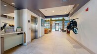 This sophisticated lobby and amenity space makes a striking first impression with its soaring double-height ceilings and expansive floor-to-ceiling steel-framed windows that flood the interior with natural light and offer views of the surrounding courtyard. Large-format light stone tile flooring extends throughout, anchoring the space with a clean, polished aesthetic.

The centerpiece of the main lounge area is a dramatic floor-to-ceiling marble fireplace, flanked by generous seating arrangements featuring tan leather sofas and classic tufted armchairs. Lush greenery and layered area rugs add warmth and texture to the otherwise contemporary design. Living moss wall accents frame the upper window panels, adding a unique organic element to the architecture.

A well-appointed bar-style counter with a striking marble waterfall surface and dark wood cabinetry serves as a focal point near the entryway, complemented by pendant lighting and bar stool seating. Nearby, a stainless steel elevator provides convenient vertical access.

A dedicated business center offers a functional retreat with high-top bar seating, a mounted flat-screen display, and a textured accent wall with warm wood framing. Additional seating areas throughout provide flexible spaces for work or socializing. The overall ambiance blends contemporary design with inviting, residential warmth.