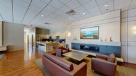 This well-appointed single-floor communal space is designed for both relaxation and gathering, blending contemporary finishes with warm, inviting elements throughout. The main lounge area serves as the centerpiece of the space, anchored by a sleek, linear gas fireplace set into a paneled accent wall. Rich hardwood flooring flows throughout, complemented by a patterned area rug beneath a seating arrangement of deep brown leather sofas and chairs. Colorful framed artwork and decorative wall sconces add character and warmth. An exposed brick accent wall near the entrance enhances the space's architectural interest, while glass entry doors allow natural light to filter through.

The fully equipped kitchen features light wood cabinetry, stainless steel appliances including a refrigerator, range, and microwave, and a striking tile mosaic backsplash. A spacious central island with bar-height seating provides an ideal spot for casual dining or socializing. Adjacent to the kitchen, a dedicated dining area offers multiple tables with chairs, bathed in natural light from large windows dressed with blinds. Recessed lighting and a suspended tile ceiling throughout ensure consistent, well-distributed illumination. Together, these zones create a cohesive, functional, and aesthetically refined communal environment.