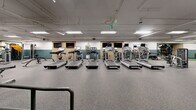 Located at 312 2nd Ave W in Seattle's Queen Anne neighborhood, this fitness center offers a generously sized, single-floor layout designed to accommodate a comprehensive range of workout activities. The space features durable speckled rubber flooring throughout, providing both safety and comfort across all exercise zones.

The cardio area is equipped with a row of treadmills, stair climbers, stationary bikes, and a rowing machine, all positioned beneath flat-screen TVs mounted along wood-paneled accent walls. Adjacent to this zone, a dedicated strength training area houses a variety of selectorized machines, a power rack with free weights, adjustable benches, and a dumbbell rack, offering a full complement of resistance training options.

A turf section with stability balls and smaller fitness accessories supports functional training and stretching. Large wall mirrors amplify the sense of space and allow users to monitor their form. Motivational phrases such as "Get Your Game On" and "You're an All Star" are displayed in bold lettering on the mirrored walls, contributing to an energetic atmosphere.

The design features a distinctive two-tone teal and white wall treatment, exposed ceiling infrastructure with fluorescent and recessed lighting, and structural columns integrated seamlessly into the layout. Handrails and accessible pathways are present, and potted plants add a subtle natural touch to the interior.