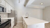 This efficient and thoughtfully designed single-floor residence offers a clean, modern aesthetic throughout. The open-concept living and kitchen area forms the heart of the unit, featuring herringbone-patterned light wood flooring that adds visual interest. The living room is bathed in natural light through large windows and is accented by crown molding, a textured ceiling, and a ceiling fan, creating a bright and airy atmosphere.

The kitchen is a functional highlight, showcasing white shaker-style cabinetry, a generous white quartz peninsula with an undermount sink and a sleek pull-down faucet, stainless steel appliances including a refrigerator and microwave, and a black electric range. Track lighting illuminates the space effectively, and vaulted ceiling details add architectural character.

The bedroom offers plush carpeting, a ceiling fan, and a window that invites natural light. A built-in closet provides practical storage. Connecting through a hallway, the bathroom is finished with white tile, a combined tub and shower, a white vanity with quartz countertop, and herringbone wood-look flooring that ties the unit together cohesively. The overall ambiance is clean, light, and move-in ready.
