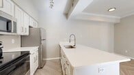 This efficient and thoughtfully designed single-floor residence offers a clean, modern aesthetic throughout. The open-concept living and kitchen area forms the heart of the unit, featuring herringbone-patterned light wood flooring that adds visual interest. The living room is bathed in natural light through large windows and is accented by crown molding, a textured ceiling, and a ceiling fan, creating a bright and airy atmosphere.

The kitchen is a functional highlight, showcasing white shaker-style cabinetry, a generous white quartz peninsula with an undermount sink and a sleek pull-down faucet, stainless steel appliances including a refrigerator and microwave, and a black electric range. Track lighting illuminates the space effectively, and vaulted ceiling details add architectural character.

The bedroom offers plush carpeting, a ceiling fan, and a window that invites natural light. A built-in closet provides practical storage. Connecting through a hallway, the bathroom is finished with white tile, a combined tub and shower, a white vanity with quartz countertop, and herringbone wood-look flooring that ties the unit together cohesively. The overall ambiance is clean, light, and move-in ready.