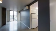 This contemporary single-floor space showcases a clean, minimalist aesthetic defined by polished concrete floors and bold slate-grey walls that create a sleek, industrial ambiance. The open-plan living area benefits from natural light streaming through a large corner window, offering views of the surrounding urban landscape and providing a bright, airy feel despite the darker color palette.

Climate comfort is handled by a wall-mounted split-system air conditioning unit, positioned high on the feature wall for efficient distribution throughout the space. The adjoining kitchen is accessed through a wide opening and is fitted with high-gloss white cabinetry extending to the ceiling, maximizing storage capacity. A full-size white refrigerator anchors the kitchen area, complementing the clean, modern finish of the cabinetry.

The overall design is cohesive and functional, with a neutral grey and white color scheme that lends itself to a variety of interior styling options. The polished concrete flooring runs seamlessly throughout, reinforcing the industrial-modern character of the space. With its efficient layout, quality finishes, and urban outlook, this space offers a refined and practical living environment well-suited to modern city living.
