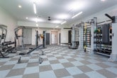 Located at 1501 Woodland Pointe Dr in Nashville, Tennessee, this single-floor fitness center offers a comprehensive workout facility designed to accommodate a wide range of exercise needs. The space is expansive and open, featuring a distinctive checkered floor tile pattern throughout that adds a clean, organized aesthetic to the environment.

The cardio zone is generously proportioned, housing a row of treadmills and multiple elliptical cross-trainers, all positioned near large windows that provide views of the surrounding outdoor greenery. Ceiling fans and suspended linear fluorescent lighting ensure the space remains comfortable and well-lit during any workout. Wall-mounted televisions are positioned throughout for entertainment during cardio sessions.

The strength training area is equally well-appointed, featuring a full rack of dumbbells paired with adjustable weight benches, a large wall mirror for form monitoring, a pull-up and dip station, and cable resistance machines. An expansive functional training wall system with resistance bands, medicine balls, and suspension trainers offers versatile exercise options. A heavy punching bag adds to the variety of available training modalities.

Additional areas include transitional corridor spaces with restroom and locker room access through classic six-panel black doors, along with seating for rest or waiting. Crown molding details lend a polished architectural finish to the facility overall.