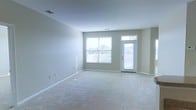 This single-floor space offers a clean, functional layout designed to maximize comfort and usability. The main living area is generously proportioned, featuring soft, wall-to-wall carpeting in a neutral tone that adds warmth underfoot while complementing the light-colored walls throughout. The room is bathed in natural light, courtesy of a large picture window fitted with horizontal blinds, allowing for flexible light control. Adjacent to the window is a private exterior door with a transom window above, further enhancing the brightness of the space and providing direct access to the outdoors. The walls are painted in a soft, neutral tone that creates a calm and welcoming atmosphere, making the space feel open and airy. Ceiling-mounted HVAC vents are discreetly integrated into the layout, ensuring year-round climate comfort. The simplicity of the design lends itself well to a variety of furnishing styles, offering a versatile canvas for personalization. Overall, the space presents a bright, well-lit, and comfortable environment ideal for everyday living.