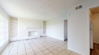 This single-story unit offers a practical and comfortable layout across its one-bedroom, one-bathroom floor plan. The living room serves as the focal point of the space, featuring a white-painted brick fireplace flanked by built-in shelving, providing both a decorative accent and functional storage. Light-colored ceramic tile flooring runs throughout the main living areas, complementing the textured ceiling and neutral white walls. A large window with vertical blinds allows natural light to fill the room, creating a bright and open atmosphere.

The kitchen is equipped with white cabinetry, a stainless steel sink, and warm-toned granite-look countertops. A square-pattern tile backsplash in earthy brown hues adds a decorative touch, and a dishwasher is included among the appliances. Wood-look flooring in the kitchen area provides a subtle contrast to the surrounding tile.

The bedroom is carpeted and features a ceiling fan with a light fixture, offering comfort and air circulation. A window brings in natural light, enhancing the room's welcoming feel. The bathroom includes a full tub and shower combination with clean white tile surround. Ceiling fans are also present in additional areas of the unit, contributing to overall ventilation and comfort throughout.
