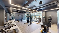 Located at 101 Washington St in Conshohocken, PA, this contemporary fitness facility spans a single floor and is thoughtfully divided into two distinct exercise rooms designed to support a wide range of workout routines. The space features an industrial-modern aesthetic, highlighted by an exposed ceiling with visible ductwork, sleek linear LED lighting, and a cool grey color palette that lends a polished, energetic atmosphere throughout.

The primary exercise room is generously sized and outfitted with a comprehensive selection of cardio equipment — including treadmills, ellipticals, and stationary bikes — arranged along expansive floor-to-ceiling windows that flood the space with natural light and offer open outdoor views. Strength training machines and free weights are also present, complemented by durable, mixed-surface flooring suited for varied workouts. A built-in media console with a flat-screen TV, decorative patterned tile accents, and a water station add functional and aesthetic detail to the room.

The second exercise room serves as a dedicated studio space, featuring warm hardwood flooring, full-wall mirrors with a ballet barre, and glass partition walls that maintain visual connectivity with the main gym floor. This versatile area is ideal for stretching, yoga, or functional training. Together, both rooms deliver a well-appointed, high-end fitness experience.