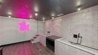 Located at 401 E Oglethorpe Ave in Savannah, Georgia, this purpose-built pet grooming space delivers a striking visual identity through bold design choices and functional layout. The interior is immediately defined by a vivid pink neon balloon dog wall sculpture, serving as a centerpiece that sets a playful yet polished tone throughout. The ceiling is finished in a deep charcoal, creating a dramatic contrast against the lighter walls and contributing to an intimate, boutique ambiance. Walls are adorned with an intricate geometric tile pattern in white and pink, lending texture and visual depth to the space. The grooming area features elevated, built-in tiled platforms with steps, each equipped with matte black handheld sprayer fixtures and open storage beneath. These stations are designed for ease of use and accessibility for both groomer and pet. Large-format tile flooring in a wood-plank style runs throughout, transitioning in tone from warm rose-brown in the main grooming area to a cooler grey near the entryway. Entry is provided through a sleek, dark-framed glass door with sidelite panel, offering a clean transition from the exterior corridor. Recessed and cage-style pendant lighting provides focused illumination across the workspace. This compact but well-appointed grooming suite is designed for efficiency and aesthetic impact.