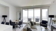This single-floor space showcases a sleek, contemporary design defined by an open-plan layout that seamlessly integrates the living and dining areas. The most striking feature is the expansive wall of floor-to-ceiling sliding glass doors, which floods the interior with abundant natural light and frames breathtaking panoramic city views from a notable elevation above the skyline.

The living area is furnished with a plush, neutral-toned sofa complemented by a dark accent armchair and a minimalist glass side table, creating a refined yet comfortable seating arrangement. Just adjacent, a compact round dining table with a pedestal base is paired with sleek, dark-framed chairs, offering an intimate dining setup that shares the same spectacular views.

The color palette throughout is restrained and sophisticated, featuring white walls, a smooth white ceiling, and light wood-toned flooring that enhances the sense of airiness. A full-length mirror adds depth to the space. The overall ambiance is calm, modern, and elevated — well-suited for those who appreciate minimalist design paired with dramatic urban vistas.