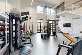 Located at 704 W Tremont Ave in Charlotte's Dilworth neighborhood, this fitness center offers a thoughtfully designed workout environment within a bright, open-plan layout. The space is defined by impressively tall ceilings that create an airy, expansive atmosphere, complemented by large multi-pane windows that flood the floor with natural light and provide views of the surrounding outdoor areas.

The main workout floor features durable, textured tile flooring and is organized to accommodate a wide variety of training styles. Strength training is well represented, with a full rack of dumbbells, a Matrix power rack with an adjustable bench and free weights, a multi-functional cable machine, and a plyo box for explosive training. Functional fitness enthusiasts will find TRX suspension trainers, resistance bands, a BOSU balance trainer, and a battle rope. A rowing machine and additional cardio equipment round out the offerings.

A wall-mounted flat-screen TV is positioned for easy viewing during workouts, with wall-mounted speakers providing audio throughout the space. A clean white staircase with modern metal railings leads to an upper-level mezzanine overlook. Glass doors open directly to an outdoor area, enhancing ventilation and connectivity to the exterior. The crisp white walls and contemporary linear lighting fixtures contribute to the space's clean, modern aesthetic.