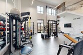 Located at 704 W Tremont Ave in Charlotte's Dilworth neighborhood, this fitness center offers a thoughtfully designed workout environment within a bright, open-plan layout. The space is defined by impressively tall ceilings that create an airy, expansive atmosphere, complemented by large multi-pane windows that flood the floor with natural light and provide views of the surrounding outdoor areas.

The main workout floor features durable, textured tile flooring and is organized to accommodate a wide variety of training styles. Strength training is well represented, with a full rack of dumbbells, a Matrix power rack with an adjustable bench and free weights, a multi-functional cable machine, and a plyo box for explosive training. Functional fitness enthusiasts will find TRX suspension trainers, resistance bands, a BOSU balance trainer, and a battle rope. A rowing machine and additional cardio equipment round out the offerings.

A wall-mounted flat-screen TV is positioned for easy viewing during workouts, with wall-mounted speakers providing audio throughout the space. A clean white staircase with modern metal railings leads to an upper-level mezzanine overlook. Glass doors open directly to an outdoor area, enhancing ventilation and connectivity to the exterior. The crisp white walls and contemporary linear lighting fixtures contribute to the space's clean, modern aesthetic.