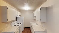 The kitchen is designed with a galley-style layout, optimizing the use of its narrow, elongated footprint. White cabinetry lines both sides of the space, providing ample upper and lower storage. The upper cabinets feature a mix of standard enclosed doors and open shelving sections, adding visual variety and accessible storage options. The countertops are finished in a speckled laminate surface that runs continuously along both sides, offering generous prep space. On one side, a white electric coil range and a full-size white refrigerator anchor the cooking area, complemented by overhead cabinetry and a ventilation unit above the stove. The opposite side features a single-basin sink with a standard faucet, set into the countertop for convenient access. The flooring is a warm-toned hardwood-style material that runs the length of the kitchen, providing a grounded contrast to the lighter cabinetry and countertops. A textured popcorn ceiling with a centrally mounted light fixture provides even overhead illumination throughout the space. The overall design is clean and utilitarian, with a neutral color palette that keeps the kitchen feeling bright and cohesive.