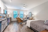 Located in Mount Pleasant, South Carolina, this single-floor studio offers a thoughtfully designed open-concept layout that seamlessly integrates living, sleeping, and dining functions into one cohesive space. Rich wood-look flooring flows throughout the main area, lending warmth and continuity to the interior.

The living and sleeping zone is generously proportioned, accommodating a full-size bed alongside comfortable seating and a sitting area. Two large windows dressed with horizontal blinds invite abundant natural light and frame views of lush surrounding greenery, creating a serene, nature-connected atmosphere. A ceiling fan adds both function and charm to this central space.

The kitchen is clean and contemporary, outfitted with crisp white shaker-style cabinetry, a subway tile backsplash, light countertops, and a full suite of stainless steel appliances including a range, microwave, and dishwasher. Light wood-look flooring in the kitchen complements the overall neutral palette.

The bathroom is finished with large-format tile flooring, a vanity with ample storage, a full mirror, and a shower-tub combination. Adjacent to the bathroom, a dedicated closet space with built-in shelving provides practical storage. The overall aesthetic throughout is calm and neutral, with soft gray and beige tones creating a relaxing, move-in-ready environment.
