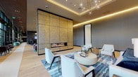 Located at 1205 Worcester St in Natick, Massachusetts, this single-floor amenity space exemplifies contemporary design with a refined, upscale aesthetic. The expansive lounge is anchored by a dramatic floor-to-ceiling gold mosaic tile fireplace wall, serving as a striking focal point that sets the tone for the entire space. Surrounding the fireplace, a carefully curated seating area features white curved armchairs, a rounded coffee table, and a richly patterned blue-and-white area rug, all resting on wide-plank light hardwood flooring. Ambient cove lighting and a sculptural globe chandelier lend warmth and elegance to the coffered ceiling above. An adjoining lounge area continues the sophisticated palette, with a white sofa, navy accent pillows, brass-accented floor lamps, and curated blue abstract artwork adorning the walls. A full-height mirror enhances the sense of depth throughout. One of the most impressive architectural features is the expansive curtain wall of floor-to-ceiling windows that lines an entire corridor, flooding the space with abundant natural light and offering sweeping views of a landscaped courtyard and the surrounding community. Softly upholstered teal swivel chairs positioned along the window wall invite relaxed seating with an outdoor connection. Throughout, the design strikes a balance between modern sophistication and welcoming comfort.