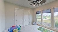 This single-floor playroom is thoughtfully designed to provide a bright, open, and functional space for children. The room is finished with large-format light-toned tile flooring that is easy to clean and maintain, lending the space a clean, modern aesthetic. A standout architectural feature is the striking windmill-style ceiling fan mounted at the center of the ceiling, adding a charming rustic-modern character to the room while ensuring comfortable airflow throughout.

Natural light fills the space generously through multiple windows fitted with adjustable roller shades, offering views of the surrounding outdoor landscape including open green spaces and a neighborhood streetscape. The abundance of windows creates a welcoming, airy atmosphere that makes the room feel open and connected to the outdoors.

The room is accessed through a set of elegant white double doors with black hardware, as well as a glass-paneled single door that provides a view into the adjoining living and dining areas, maintaining a sense of openness and visual connectivity with the rest of the home. The neutral, light-gray wall tones throughout provide a versatile backdrop that complements a wide range of uses and décor. Ample floor space allows for flexible arrangement of play areas, making this room a practical and inviting retreat for children within the home.