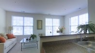 This single-floor space offers a clean, open layout bathed in natural light streaming through multiple large windows fitted with horizontal blinds. The generous fenestration creates an airy, inviting atmosphere while offering views of the surrounding exterior. A prominent granite-topped counter or bar serves as a natural dividing element within the open floor plan, adding a touch of elegance with its richly patterned stone surface. The living area features a sleek glass-topped coffee table with a classic iron frame, complemented by decorative touches such as a framed botanical artwork and a vibrant arrangement of orange flowers that bring warmth and color to the neutral palette. The walls are finished in a crisp, light tone that enhances the sense of openness, while the smooth ceilings maintain a clean, uncluttered look. Lush green potted plants are thoughtfully placed throughout, adding a natural element to the interior. The overall ambiance is calm and well-organized, with a balanced combination of functional surfaces and decorative accents that make the space both practical and aesthetically pleasing.