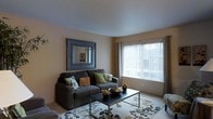 This single-floor residence offers a thoughtfully arranged living space designed for comfort and functionality. The living room serves as the central gathering area, finished in warm neutral tones that create an inviting and relaxed atmosphere throughout. A large picture window with vertical blinds and decorative curtain panels allows generous natural light to fill the room, lending the space an open, airy feel. The smooth, clean ceiling enhances the sense of openness, while the soft carpeted flooring adds warmth underfoot. A plush sofa anchors the seating arrangement, complemented by a patterned area rug that adds visual interest to the floor. A sleek coffee table provides a functional centerpiece to the living area. A framed wall mirror adds depth and dimension to the main wall, reflecting light and contributing to the overall brightness of the space. The layout flows naturally, making efficient use of the available footprint while maintaining a sense of spaciousness. Overall, this single-level space presents a clean, comfortable, and well-lit environment with a neutral palette that provides a versatile backdrop for a variety of interior styles.
