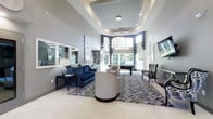 This elegantly appointed single-floor space exudes a refined, upscale ambiance with a seamless blend of comfort and sophistication. The centerpiece of the main lounge area is its dramatically tall, coffered ceiling with recessed lighting and a modern chandelier, lending a grand sense of openness and volume to the interior. Large arched windows at the far end flood the space with natural light, offering views of lush greenery outside and creating a bright, welcoming atmosphere throughout.

The seating area is thoughtfully arranged with a rich navy blue velvet sofa, neutral swivel accent chairs, and patterned armchairs, all anchored by an ornate area rug in a complementary blue-and-white palette. The light-toned large-format tile flooring adds a clean, polished finish underfoot. Mirrored wall panels and framed artwork contribute to the space's stylish, contemporary aesthetic.

Beyond the main lounge, a billiards table is positioned near the windows, making it an ideal recreational focal point. The space is further enhanced by soft layered lighting, including pendant fixtures and recessed downlights, ensuring a warm and inviting environment at any time of day. Overall, this space delivers a high-end, functional setting well-suited for socializing and leisure.