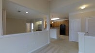 This single-floor residence offers an open and airy layout that connects the main living area to the kitchen in a fluid, functional design. The living space features neutral carpeting and crisp white walls, creating a clean and welcoming atmosphere. A half-wall partition with a pass-through opening visually connects the living area to an adjacent room, which features built-in shelving — ideal for storage or display.

The kitchen is a standout feature, outfitted with warm honey-toned wood cabinetry that extends to the upper walls, providing generous storage. A center island anchors the kitchen space and offers additional prep and seating potential. Black appliances, including a full-size refrigerator, add a bold contrast to the lighter cabinetry and countertops. Overhead pendant lighting illuminates the kitchen, complementing the recessed lighting found throughout the space.

The flooring transitions from plush carpet in the living areas to smooth tile in the kitchen, clearly defining each zone while maintaining a cohesive aesthetic. A private door off the kitchen leads to an additional room, adding versatility to the overall layout. High ceilings contribute to a sense of openness and volume throughout this well-appointed, single-level home.
