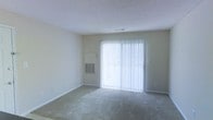 This single-floor space offers a clean and functional layout designed for comfortable everyday living. The main living area is a standout feature, bathed in natural light streaming through a large sliding glass door fitted with vertical blinds, creating a bright and airy atmosphere. The room is finished with wall-to-wall carpeting, providing a soft and comfortable underfoot feel throughout. A textured popcorn ceiling adds a classic touch to the interior, while neutral-toned walls keep the space feeling open and versatile, ready to accommodate a variety of furnishing styles. A wall-mounted HVAC unit ensures year-round climate control directly within the living space. The sliding glass door not only brings in abundant natural light but also provides convenient access to the outdoors. Overall, this space presents a straightforward, practical interior with a light-filled ambiance, neutral finishes, and essential amenities that make it a functional and adaptable environment.