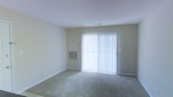 This single-floor space offers a clean and functional layout designed for comfortable everyday living. The main living area is a standout feature, bathed in natural light streaming through a large sliding glass door fitted with vertical blinds, creating a bright and airy atmosphere. The room is finished with wall-to-wall carpeting, providing a soft and comfortable underfoot feel throughout. A textured popcorn ceiling adds a classic touch to the interior, while neutral-toned walls keep the space feeling open and versatile, ready to accommodate a variety of furnishing styles. A wall-mounted HVAC unit ensures year-round climate control directly within the living space. The sliding glass door not only brings in abundant natural light but also provides convenient access to the outdoors. Overall, this space presents a straightforward, practical interior with a light-filled ambiance, neutral finishes, and essential amenities that make it a functional and adaptable environment.
