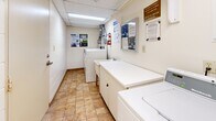 Located at 234 Oakdale Ave in St. Catharines, Ontario, this shared laundry room is a well-organized, single-floor utility space designed to serve building residents efficiently. The room features a compact yet functional layout, with multiple Huebsch top-load washing machines and matching dryers arranged along the walls, providing ample capacity for shared use. A deep utility sink sits near the entrance, offering a convenient handwashing or pre-treatment station. White laminate folding counters with cabinet storage below run along one side of the room, providing a practical surface for sorting and folding laundry. A hot water tank is housed at the rear of the room alongside a louvered utility closet, keeping mechanical components neatly contained. The floor is finished in earth-toned ceramic tile, contributing to a clean and easy-to-maintain environment. Overhead fluorescent lighting ensures the space is well-lit throughout. The walls are lined with informational notices, usage guidelines, and building communications boards, keeping residents informed. A fire extinguisher is mounted on the wall, and ventilation ductwork for the dryers is visible overhead. The room is maintained with a clear emphasis on cleanliness, safety, and resident convenience.