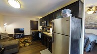 This single-floor residence offers a cohesive and functional layout designed for comfortable everyday living. The kitchen serves as a central highlight, showcasing rich, dark espresso cabinetry that extends from floor to ceiling, providing generous storage space. Stainless steel appliances, including a full-size refrigerator, a gas range, and an over-the-range microwave, lend a modern and polished look to the space. The countertops feature a warm, neutral tone that complements the bold cabinetry, while a classic tile backsplash adds a subtle decorative touch. Dark hardwood flooring runs throughout, creating a sense of continuity and warmth across the living areas. A patterned area rug near the living space adds texture and visual interest. The kitchen flows naturally into the adjacent living area, making the overall layout feel open and connected. A textured ceiling adds a traditional character to the interior. The entry door visible in the background is finished in a rich wood tone, reinforcing the warm, cohesive design palette found throughout the space. Overall, the interior presents a well-maintained and thoughtfully designed environment suited for functional, day-to-day living.