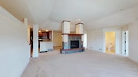 This single-floor home showcases a thoughtfully designed open-concept layout that maximizes both space and functionality. The living area is anchored by a commanding central fireplace clad in slate tile with rich wood trim accents and a raised hearth, serving as the visual focal point of the room. Twin white columns flank the fireplace surround, adding a sense of architectural structure and elegance. Soaring vaulted ceilings in the living space create an airy, expansive atmosphere, enhanced by smooth, neutral-toned carpet that flows throughout. The kitchen is positioned on a slightly elevated level and features warm cherry wood cabinetry, a peninsula with countertop seating, and black appliances, offering both style and practicality. A half-wall and open passthrough connect the kitchen to the living area, maintaining the open feel while subtly defining each space. Recessed and flush-mount lighting provide warm, even illumination throughout. A doorway visible in the background hints at additional rooms beyond the main living space. Overall, the home exudes a warm, welcoming ambiance with a blend of classic and contemporary design elements.