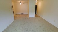 This single-floor space offers an open and functional layout with a straightforward, practical design. The main living area is carpeted throughout, providing a soft underfoot feel across a generously open floor plan. A ceiling fan with an integrated light fixture is centrally positioned, contributing to both air circulation and ambient lighting in the space.

A structural column serves as a natural divider within the room, subtly separating the space into distinct zones without fully closing off the layout — maintaining an open, connected feel. The walls are finished in a clean, neutral white tone, lending the interior a bright and versatile backdrop suitable for a variety of furnishing styles.

A baseboard heater is visible along one wall, indicating a reliable built-in heating source. The doorways visible from the main area lead to additional rooms, suggesting a functional flow throughout the unit. Overall, the space presents a clean, open canvas with practical features, well-suited for comfortable everyday living.
