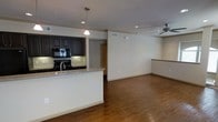 This single-floor space showcases a thoughtfully designed open-concept layout that seamlessly connects the kitchen and living areas. The kitchen is a standout feature, boasting rich dark cabinetry, granite countertops, and a peninsula with a breakfast bar that doubles as a natural divider between the cooking and living spaces. A pendant light hangs above the peninsula, adding a focused point of illumination, while recessed lighting throughout ensures the space remains bright and welcoming. Built-in black appliances, including a microwave and stove, are neatly integrated into the cabinetry, and a tile backsplash adds a refined finishing touch. The living area is generously proportioned, with warm hardwood flooring running throughout that adds both character and continuity. A ceiling fan with integrated lighting anchors the living space, providing both comfort and ambient light. The neutral white walls create a clean, versatile backdrop, while crown molding along the ceiling adds a subtle architectural detail. A secondary counter area or bar is visible along one wall, offering additional functionality. The overall ambiance is modern, clean, and open, making the space feel inviting and highly livable.
