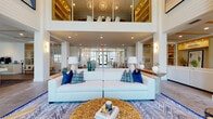 Located at 1650 Clearwater Largo Rd in Largo, Florida, this single-story community amenity building delivers an impressive coastal-modern aesthetic from the moment you enter. The grand lobby commands attention with its soaring double-height ceilings, floor-to-ceiling black-framed windows, and an abundance of natural light that floods the open interior. A curated seating area anchors the space, featuring a plush white sectional sofa accented with deep navy and teal throw pillows, wood-framed accent chairs, and a striking natural wood slice coffee table atop a bold geometric area rug — all contributing to the refined, beach-inspired atmosphere. A mezzanine level is visible overhead, adding vertical drama to the central gathering area. Adjacent to the lobby, a sleek reception and service corridor features large-format tile flooring, pendant lighting, and a long white counter flanked by glass-partitioned private rooms with modern framed doors. A dedicated conference or meeting room offers an oval table, upholstered seating, and views of lush tropical landscaping through wide windows. The leasing or administrative area showcases decorative blue encaustic tile, built-in wood shelving, and contemporary cabinetry. Throughout, the design balances clean, white architectural lines with warm wood accents and coastal color palettes.