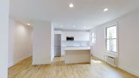 This single-floor space showcases a clean, contemporary design with an open-concept layout that seamlessly connects the kitchen and living areas. The kitchen is a standout feature, outfitted with floor-to-ceiling white cabinetry that maximizes storage while maintaining a streamlined, minimalist aesthetic. A central island provides additional workspace and serves as a natural gathering point. Stainless steel appliances, including a full-size refrigerator and built-in microwave, complement the modern design. Recessed lighting is installed throughout the ceiling, providing even, adjustable illumination across the space. Light-toned hardwood flooring runs continuously throughout, enhancing the sense of openness and warmth. Natural light enters through double-hung windows, contributing to the bright, airy atmosphere. The white walls and white trim maintain a cohesive, neutral palette that allows the architectural details and finishes to speak for themselves. The overall ambiance is fresh, clean, and functional — well-suited for those who appreciate modern design and efficient use of space. The open floor plan offers flexibility in furniture arrangement, making the living area highly adaptable to various lifestyle needs.